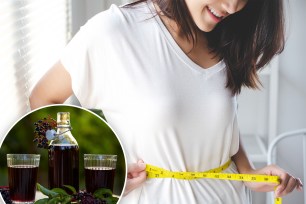 A woman smiles while measuring her waist. A glass of elderberry juice sits on a table.